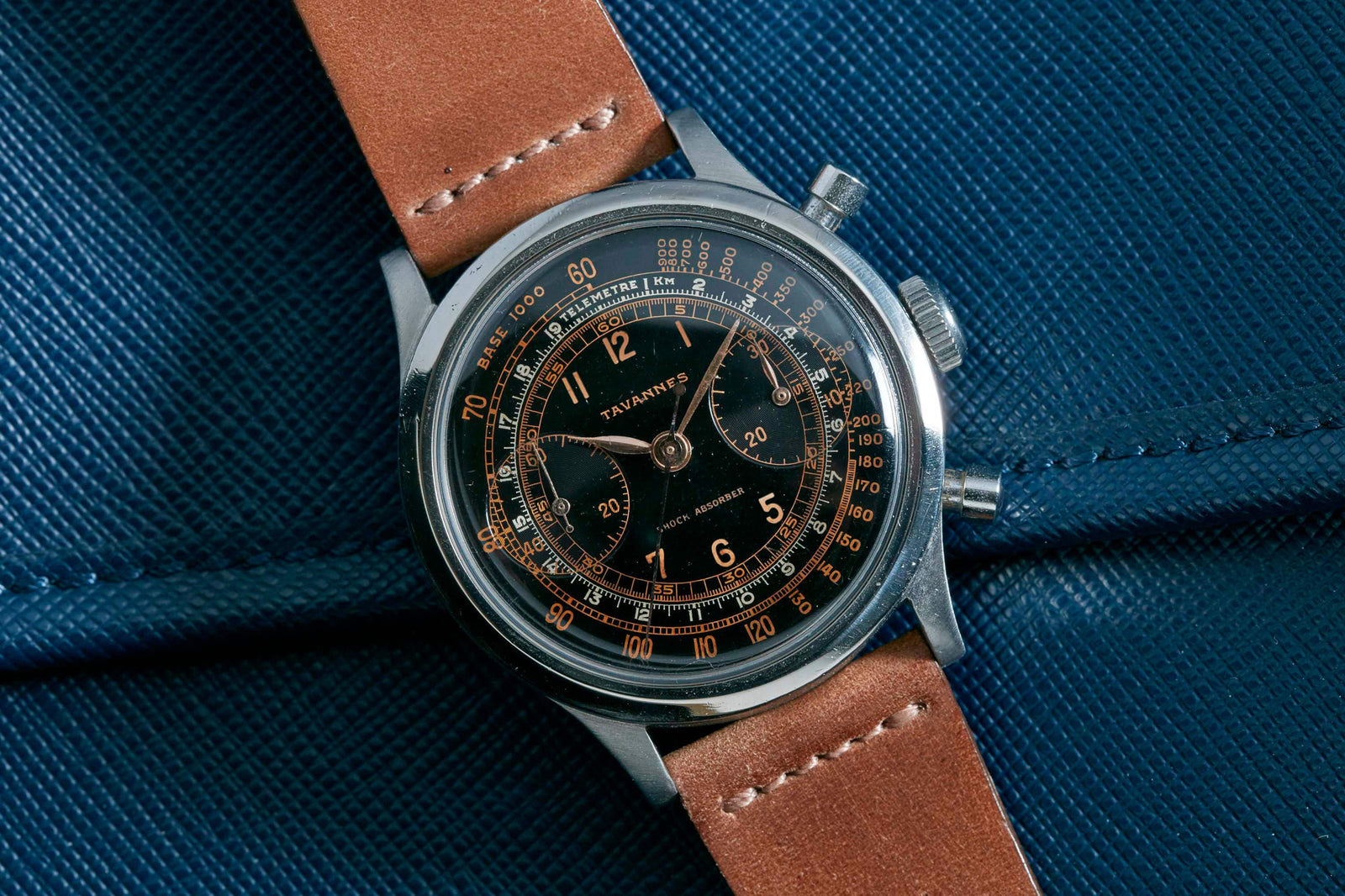 The Tavannes Chronograph, a vintage-style wristwatch with a brown leather strap and black dial featuring gold numerals, displays the Tavannes name on its face as it rests on a textured dark blue surface.