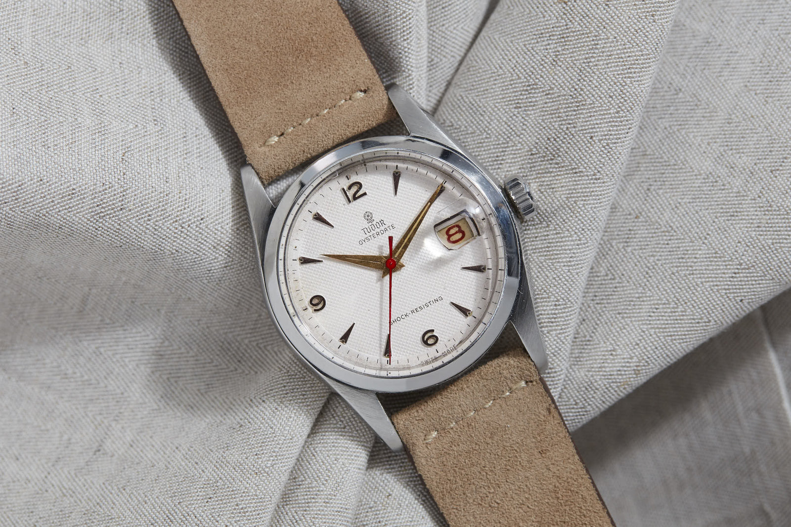 The Tudor Oyster Prince by Tudor features a silver case, textured white dial, tan suede strap, and red second hand. Inspired by the Rolex Oyster case, it has automatic movement and bold Arabic numerals at 3, 6, 9, and 12.