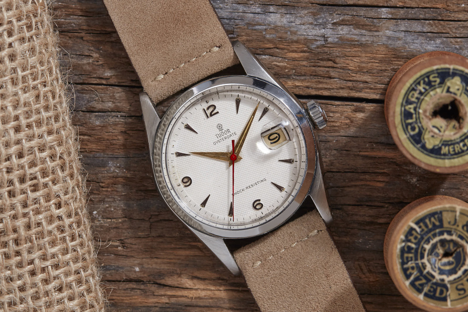 A classic Tudor Oyster Prince watch by Tudor, paired with a beige suede strap and the renowned Rolex Oyster case, lies on rustic wood beside burlap and vintage wooden thread spools, showing the date as the 9th.