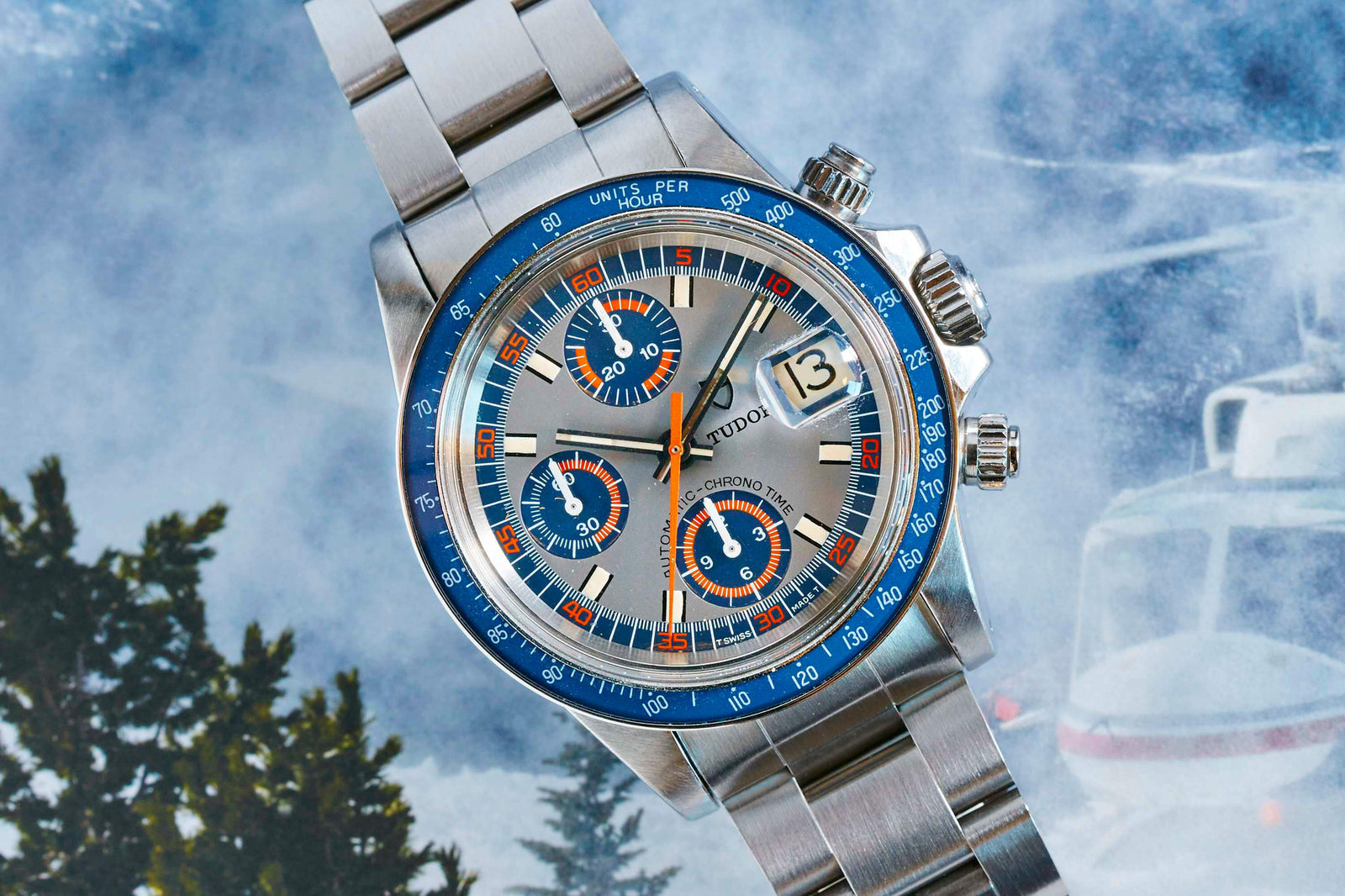 The Tudor Monte Carlo 9420/0, a stainless steel chronograph with blue and orange bezel and subdials on a metal bracelet, is shown in a snowy scene with a helicopter and trees—capturing the vintage charm of Tudor watches.