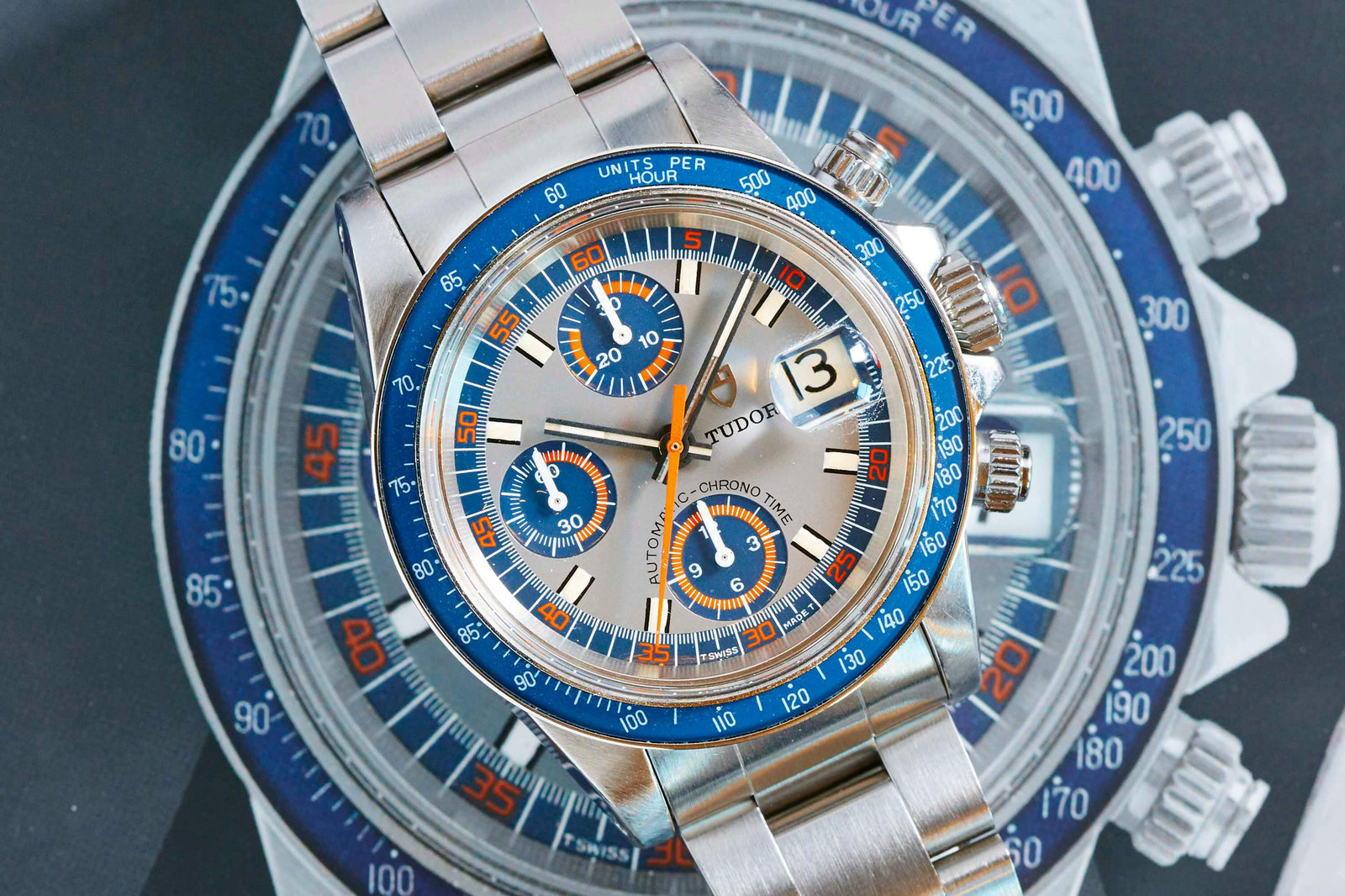 A Tudor Monte Carlo 9420/0 chronograph watch with a blue and orange bezel, silver dial, and metal bracelet rests atop a similar watch image, highlighting the refined vintage details of Tudor craftsmanship.