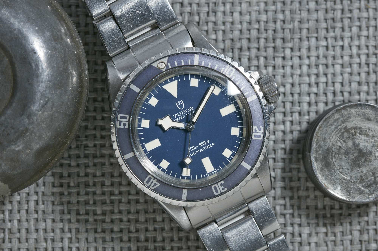 A Tudor Submariner Snowflake stainless steel watch with a blue dial and bezel is displayed on a woven gray surface, alongside part of a metal object. Detailed description coming soon!.