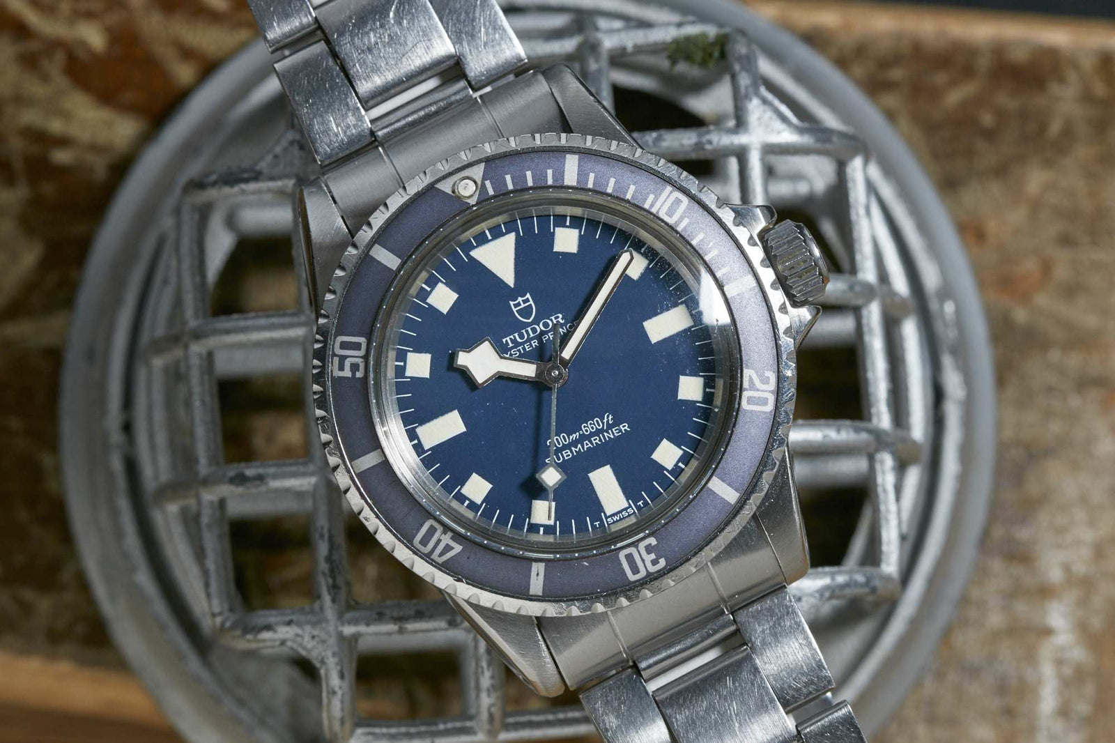 A Tudor Submariner Snowflake wristwatch by Tudor, featuring a blue dial and bezel, silver hour markers, and a stainless steel bracelet, rests on a metallic grated surface. Detailed description coming soon!.