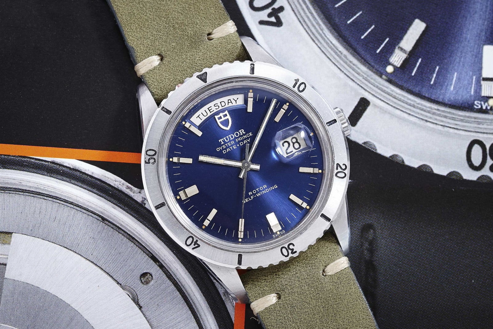 The Tudor Date-Day 7020/0 Blue Dial by Tudor displays 2:38 and Tuesday the 28th, featuring a silver bezel, white markers, and an olive-green strap, set against a backdrop of watch images.