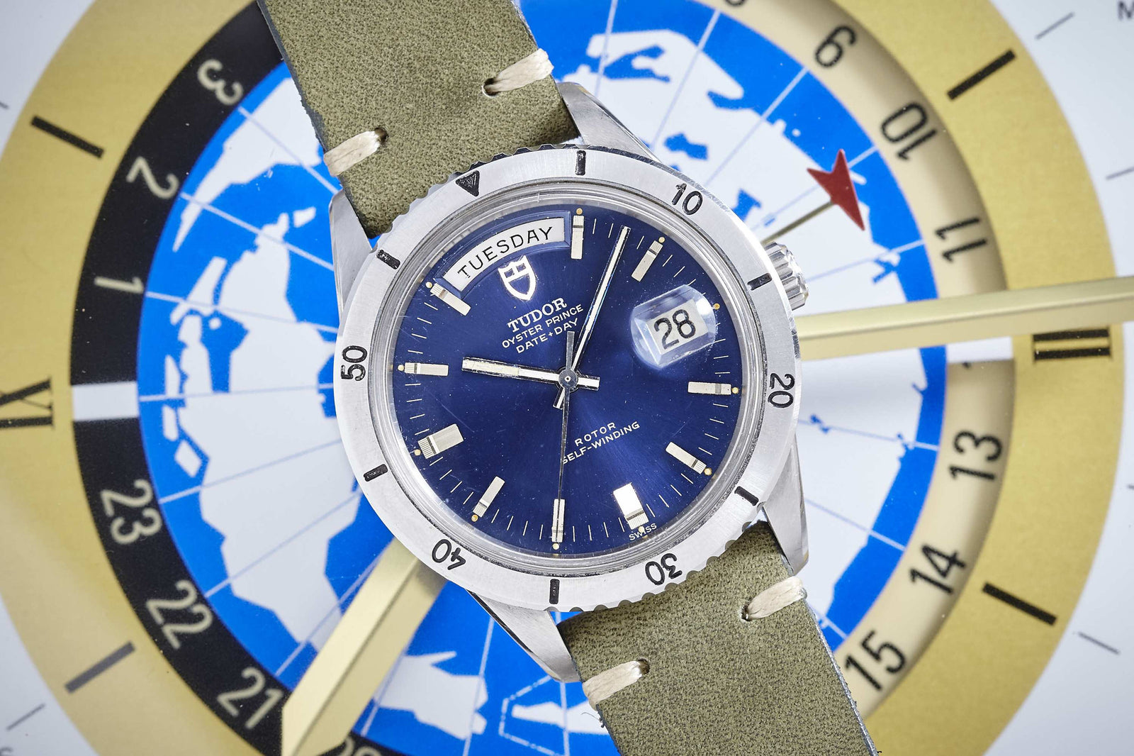 A Tudor Date-Day 7020/0 Blue Dial wristwatch with an olive-green strap and day-date display is set on a globe-themed clock face featuring gold and blue accents and a prominent gold hand.