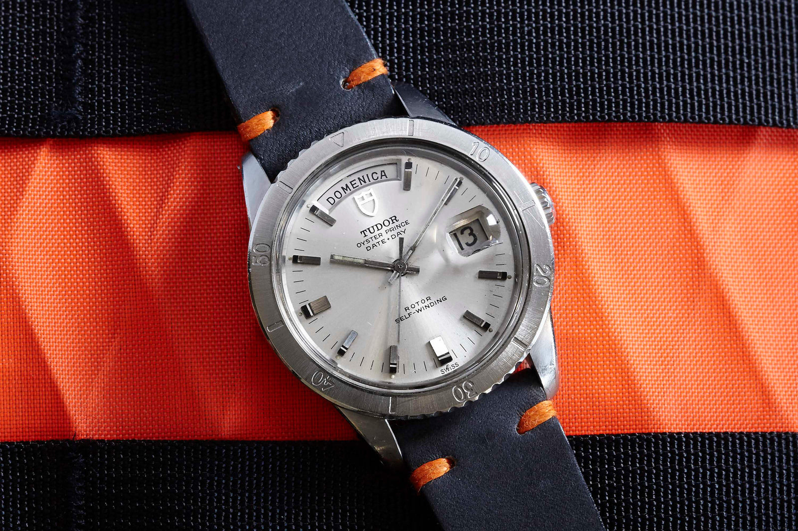 A Tudor Date+Day wristwatch by Tudor sits on blue and orange striped fabric, featuring a white dial with black hour markers, date window, and a bold black strap accented with orange stitching.