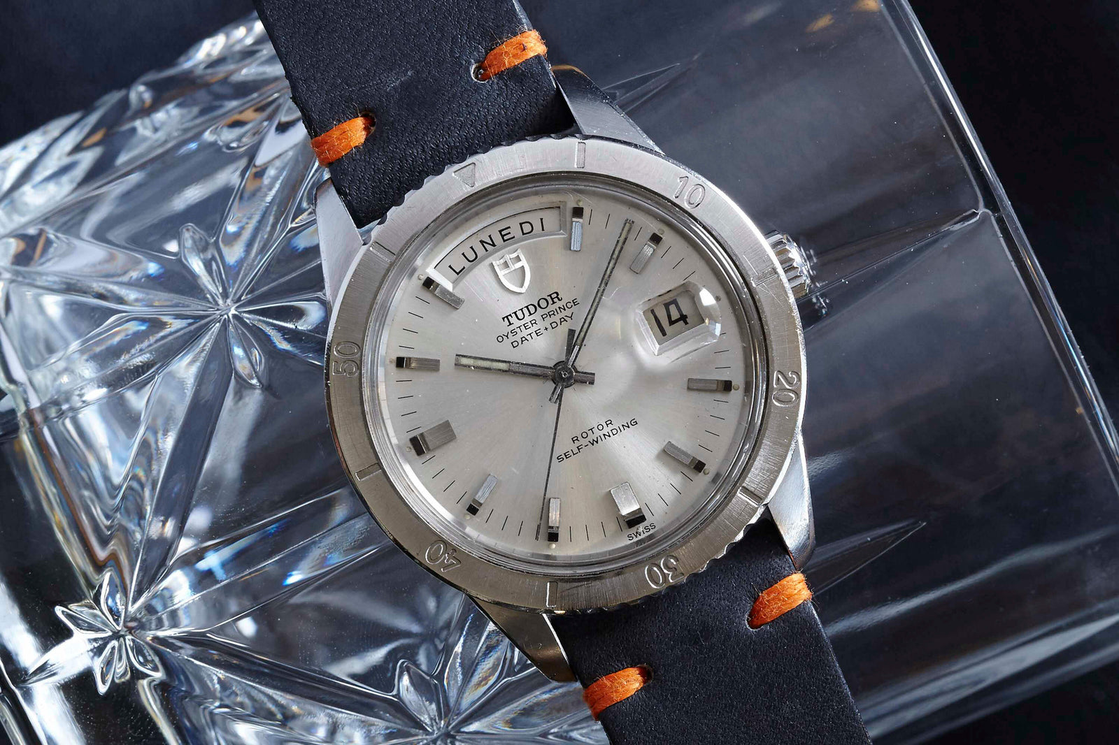 A Tudor Date+Day by Tudor with a silver case and black leather strap with orange stitching, showing LUNEDÌ and the date 19, rests elegantly on a patterned glass surface.