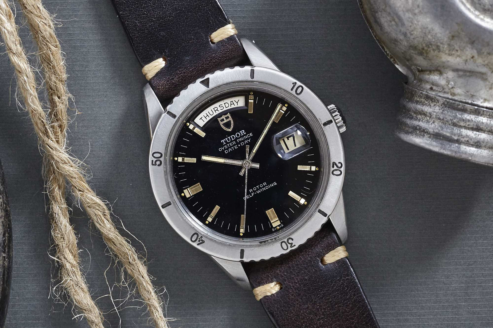 A Tudor Date-Day wristwatch by Tudor, featuring a black dial, day and date windows, silver bezel, and brown leather strap, is displayed on a gray surface next to rope and a metal object.