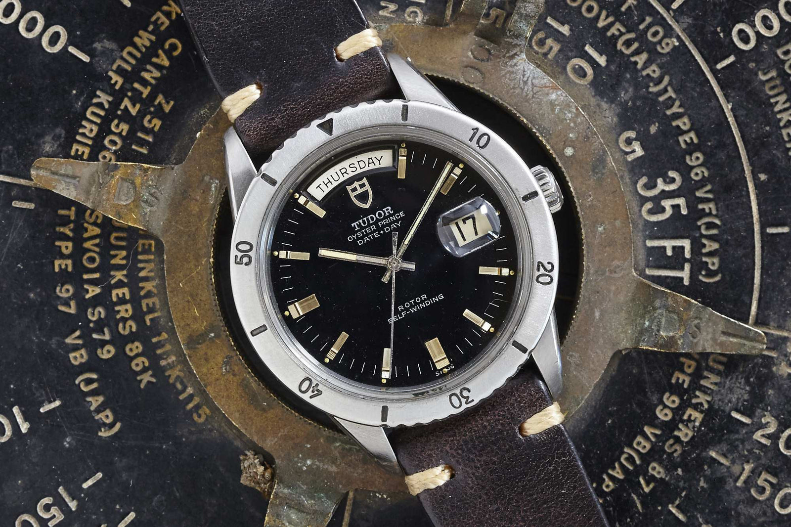 A vintage Tudor Date-Day by Tudor with black dial and brown leather strap features day, date, and silver bezel. Styled like a classic steel Rolex Day-Date, it's displayed on an old metal gauge for a timeless look.