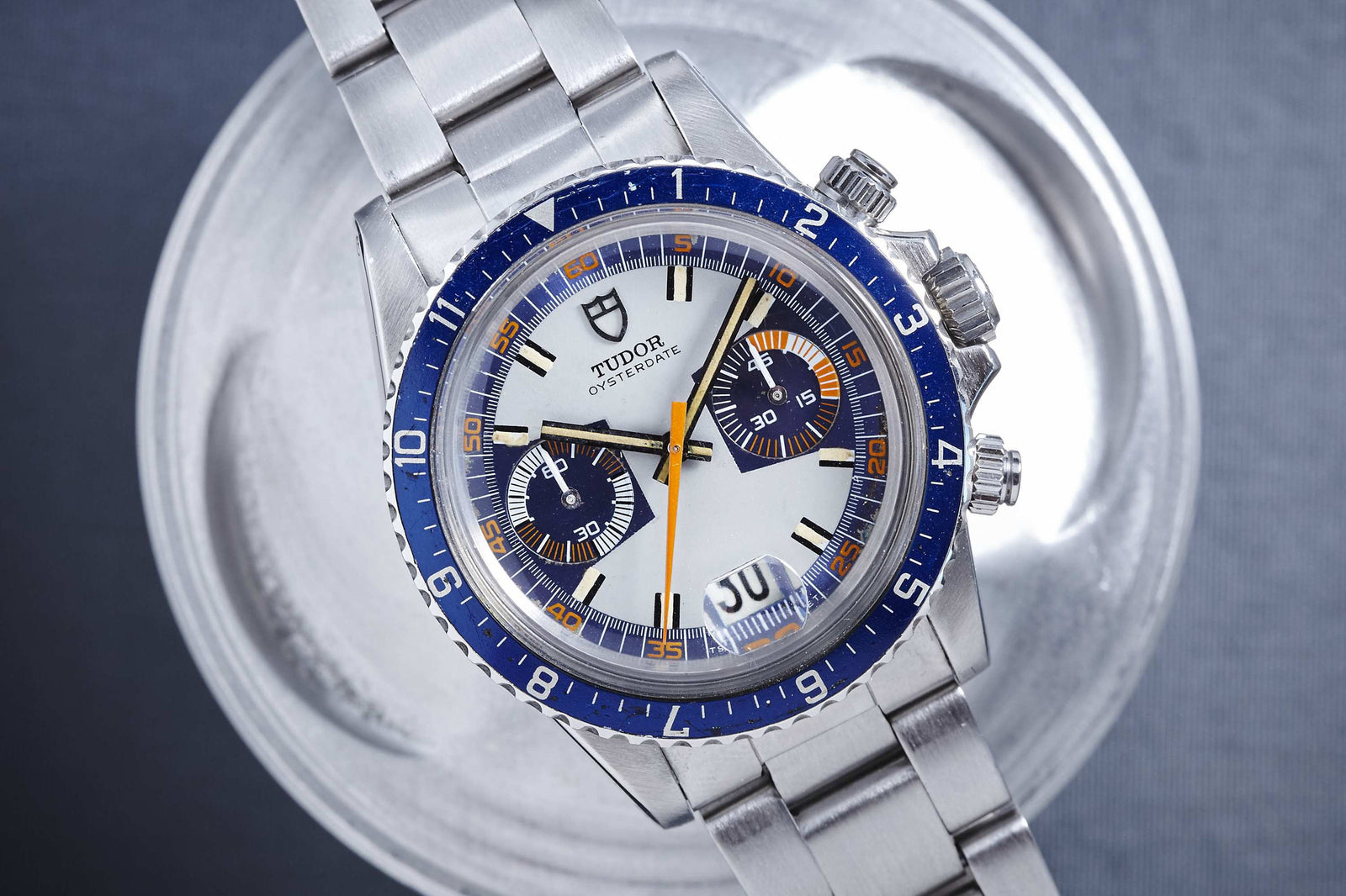 A close-up of the Tudor Monte Carlo by Tudor, highlighting its blue bezel, white dial, stainless steel bracelet, and two subdials with date display as it rests on a round metallic surface.