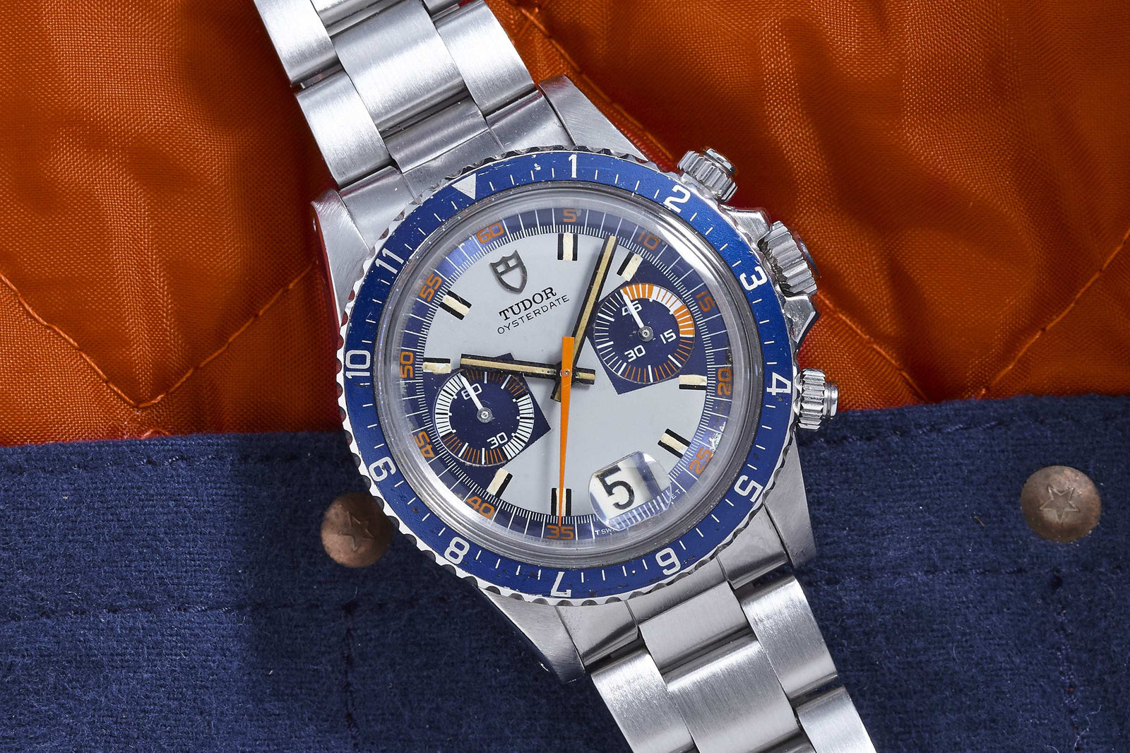 The Tudor Monte Carlo by Tudor features a stainless steel case, blue and orange bezel, white dial with two subdials—all styled against an orange and blue fabric with metal rivets—an iconic vintage chronograph wristwatch.