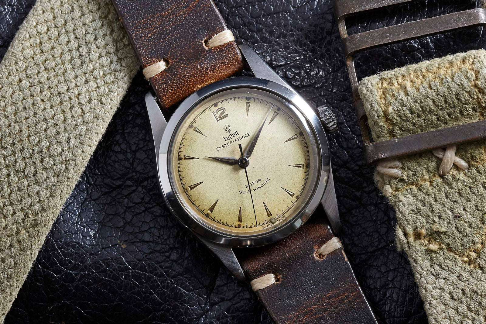 A Tudor Oyster Prince wristwatch by Tudor, featuring a cream dial, brown leather strap, and silver casing, is displayed on a textured black surface next to two pieces of worn beige fabric.
