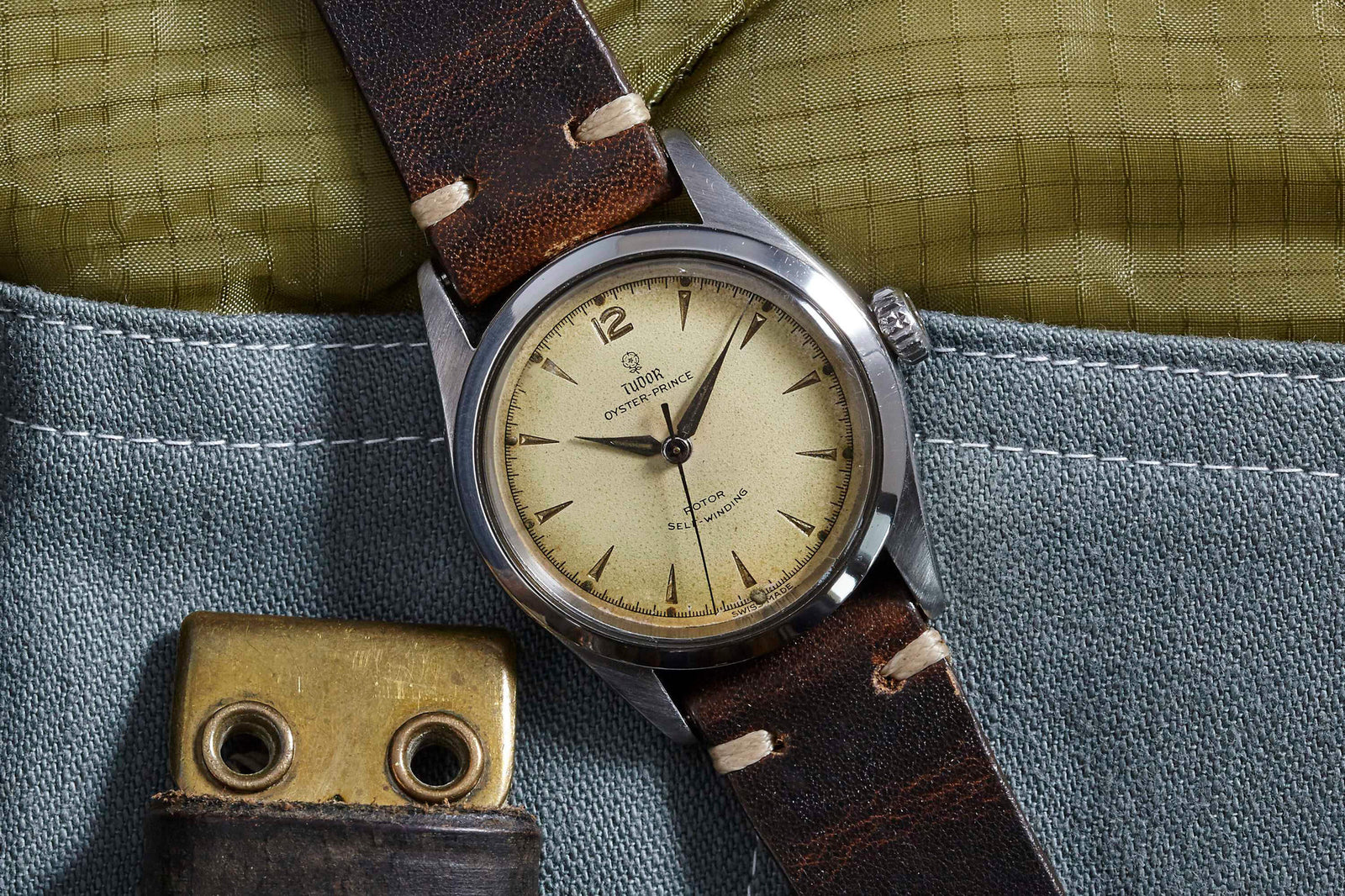 A Tudor Oyster Prince wristwatch by Tudor, featuring a cream dial and leather strap, rests on textured green and blue fabric beside a brass snap button, evoking the enduring legacy of classic tool watches.