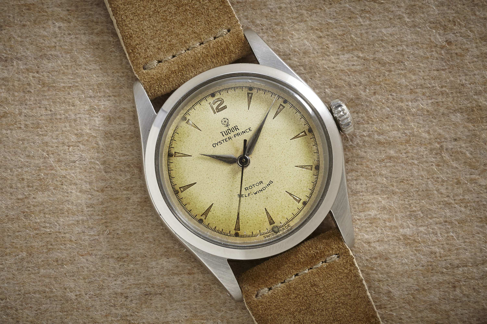 The Tudor Oyster Prince by Tudor features a vintage stainless steel case, cream dial with silver markers, and a brown suede strap, showcased on a textured beige surface.