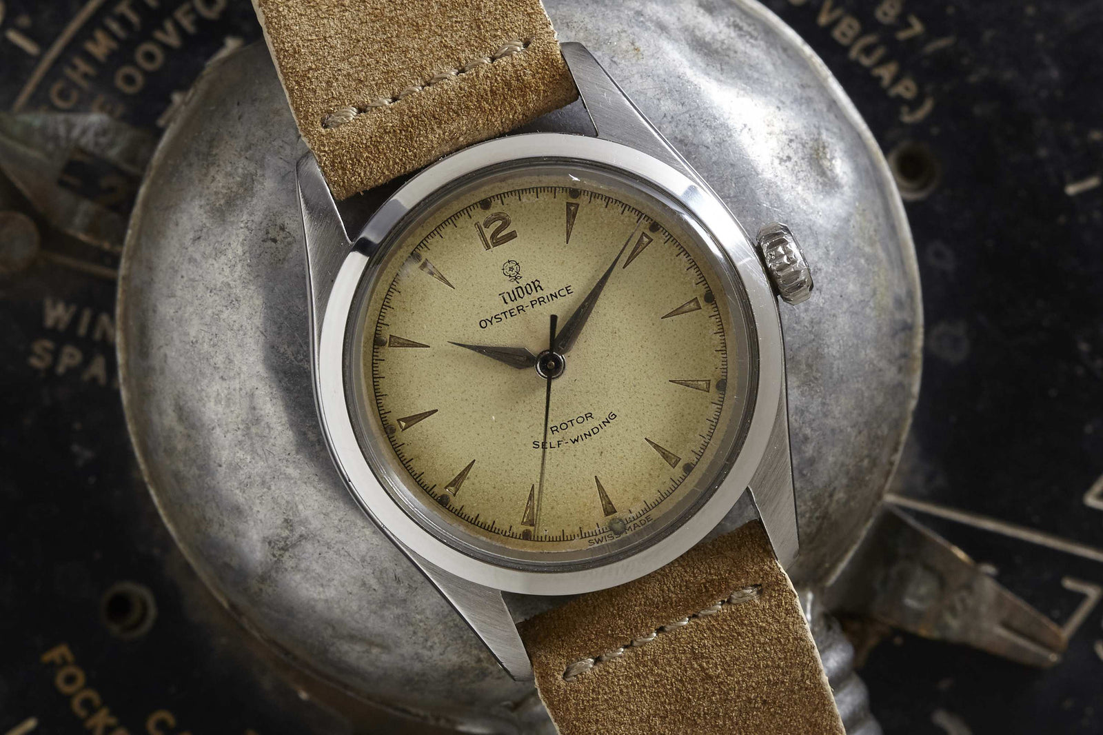 A Tudor Oyster Prince by Tudor, featuring a vintage stainless steel case, tan suede strap, and faded beige dial, rests on an industrial metallic surface with dark, worn textures in the background.