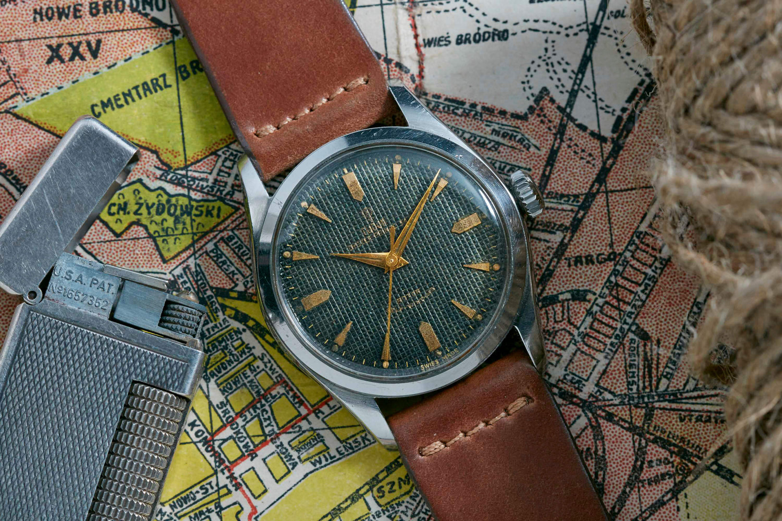 A Tudor Oyster Prince Waffle Dial wristwatch with a brown leather strap rests on an old map, accompanied by a metal lighter and rope. Its textured greenish dial, gold markers, and Swiss ETA movement evoke classic Tudor style.