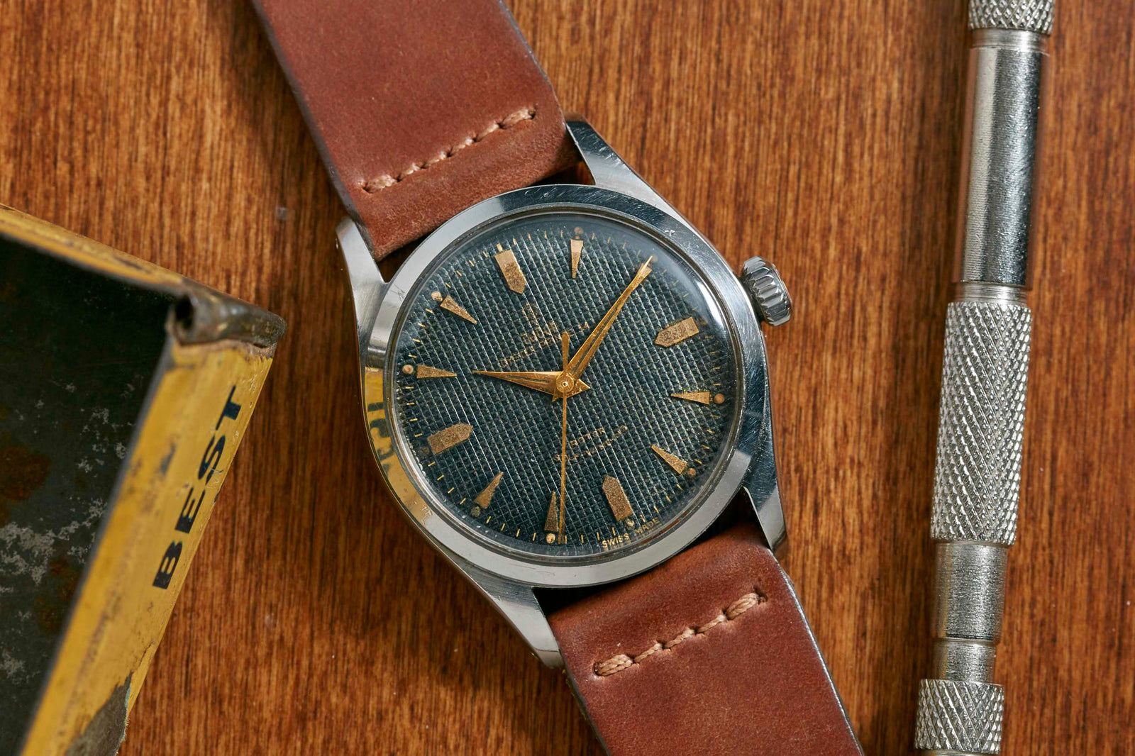 The Tudor Oyster Prince Waffle Dial by Tudor features a textured dark dial, gold indices, and a brown leather strap. Resting on wood near a metal tool and yellow box, it boasts reliable ETA Swiss movements.
