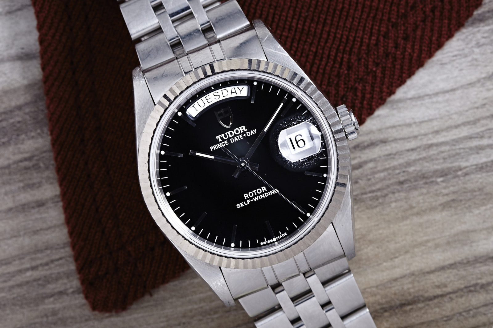 A Tudor Prince Date-Day Black Dial watch by Tudor, featuring a 36mm steel case, displays TUESDAY and the date 16. It rests on a wooden surface with a textured dark brown cloth underneath.