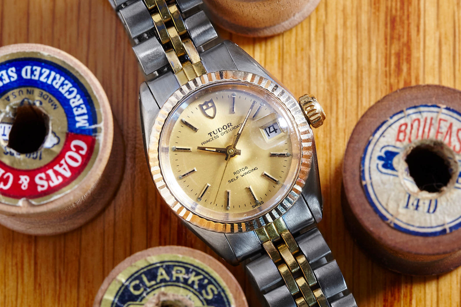 A Dreffa Tudor Princess Oysterdate Two Tone wristwatch with a gold and silver band rests on a wooden surface among vintage sewing thread spools. The watch features a date display, gold hour markers, and matching hands. Description coming soon!.
