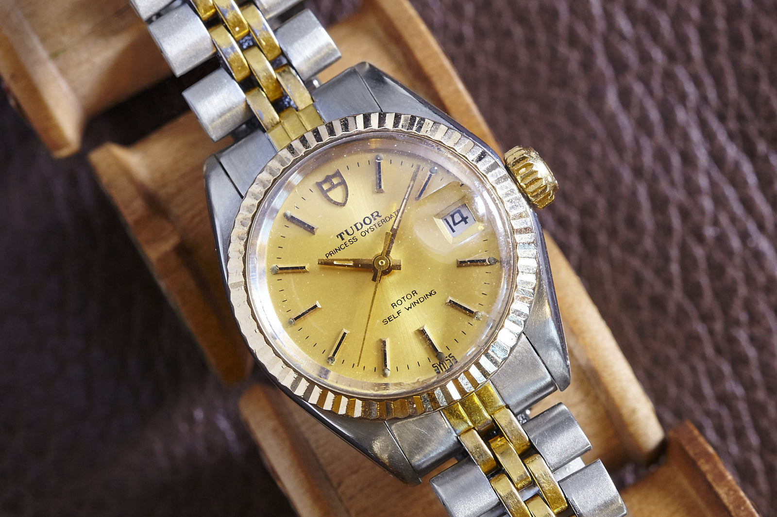 A close-up of a Dreffa Tudor Princess Oysterdate Two Tone watch with a gold and silver band, gold dial, and date window showing the 14th, displayed on a wooden watch holder. Description coming soon!.