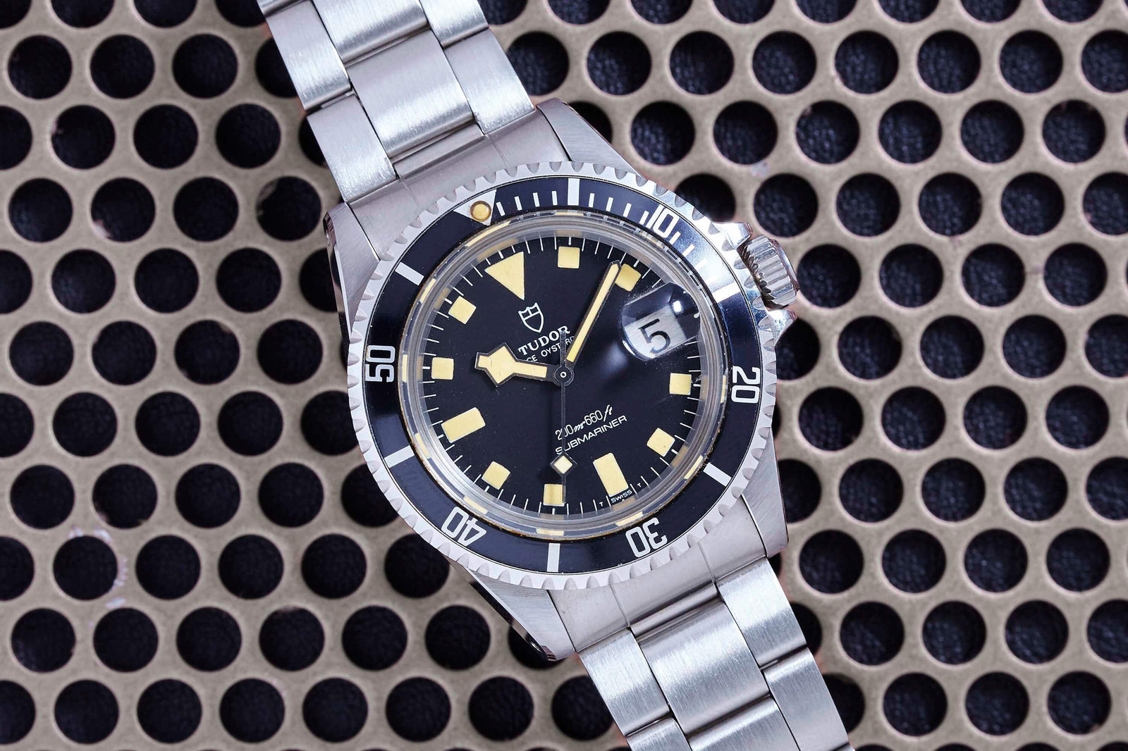 A Tudor Snowflake Submariner stainless steel wristwatch with a black dial, yellow hour markers, and a date window is shown on a silver metal bracelet, resting on a perforated metal surface.