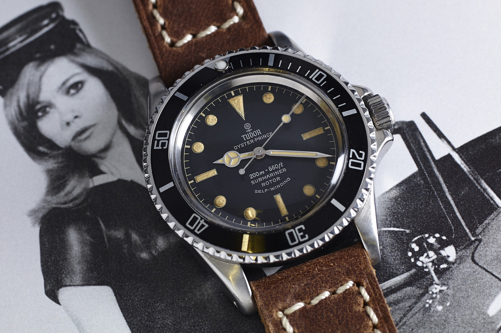A Tudor Submariner 7928 Gilt Dial with a brown leather strap lies atop a black-and-white photo of a woman in a hat beside a classic car.