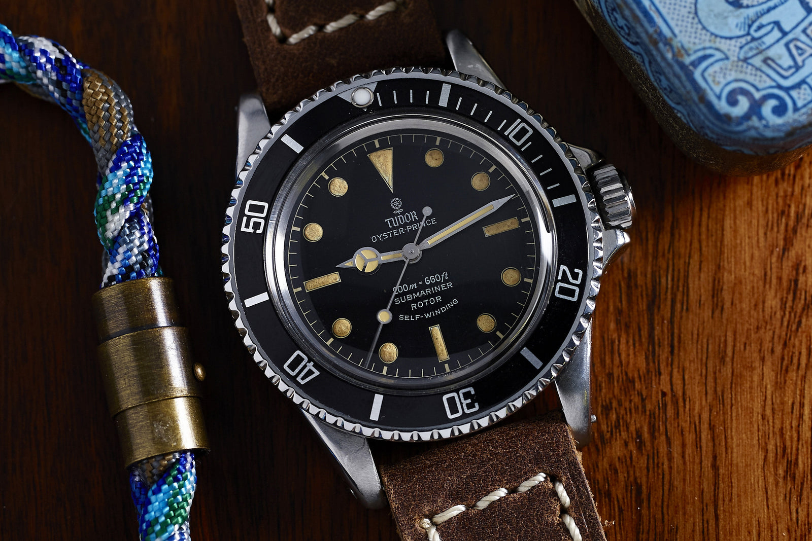 A Tudor Submariner 7928 Gilt Dial watch by Tudor with a gilt dial and brown leather strap sits on a wooden surface next to a blue object and a colorful braided bracelet.