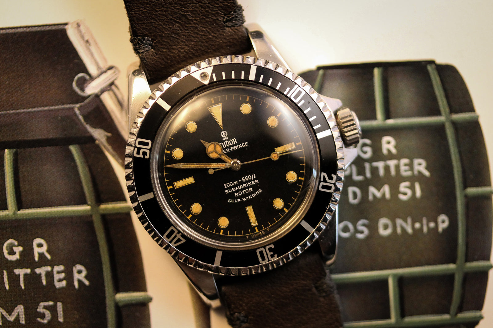 A close-up of the Tudor Submariner 7928 Gilt Dial PCG wristwatch with a black bezel and leather strap, displayed on military-themed items featuring GR GLITTER DM 51 text.