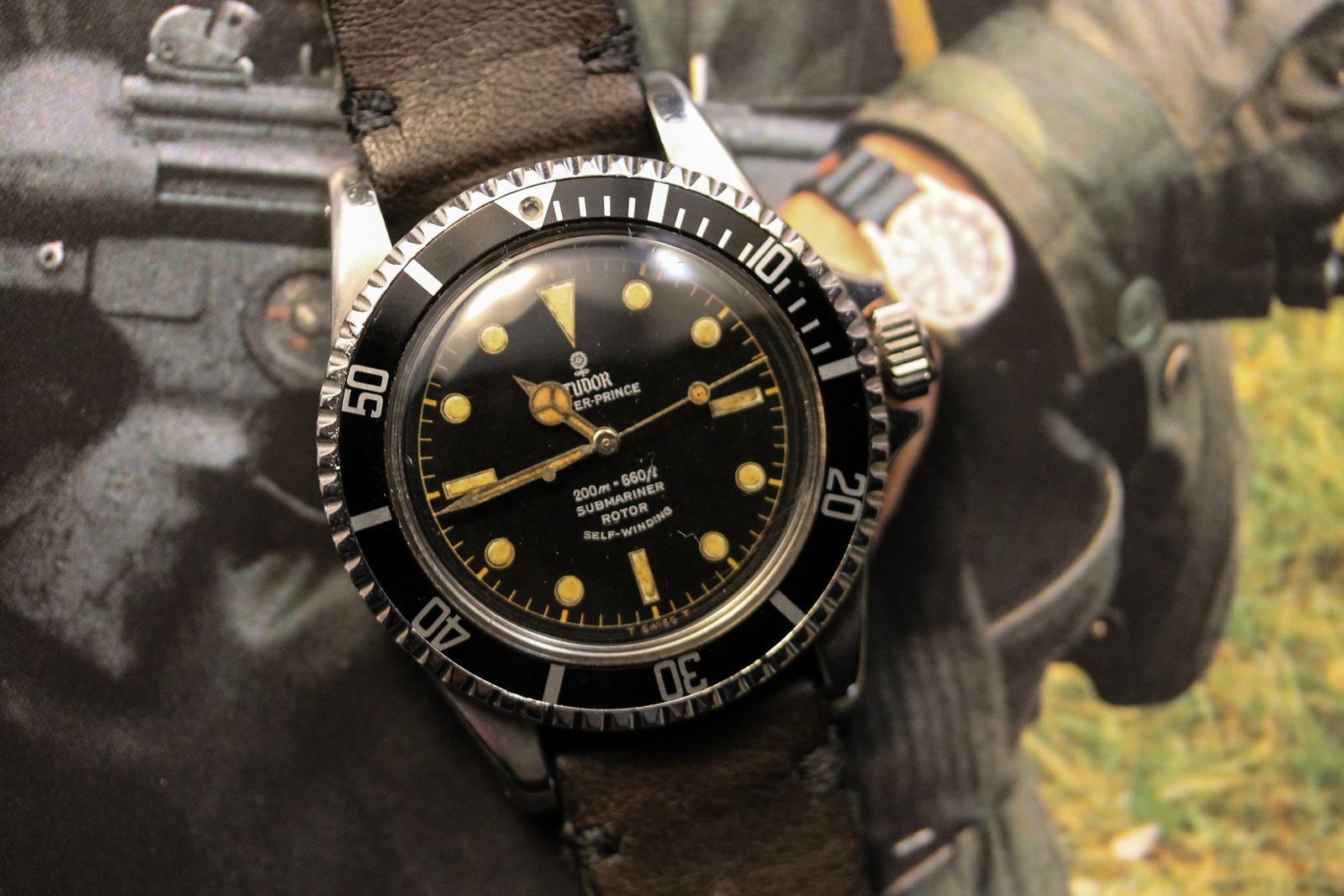 A close-up of a vintage Tudor Submariner 7928 Gilt Dial PCG by Tudor, featuring a black dial with luminous markers and a brown leather strap, set before a blurred background of someone wearing a watch while handling equipment.