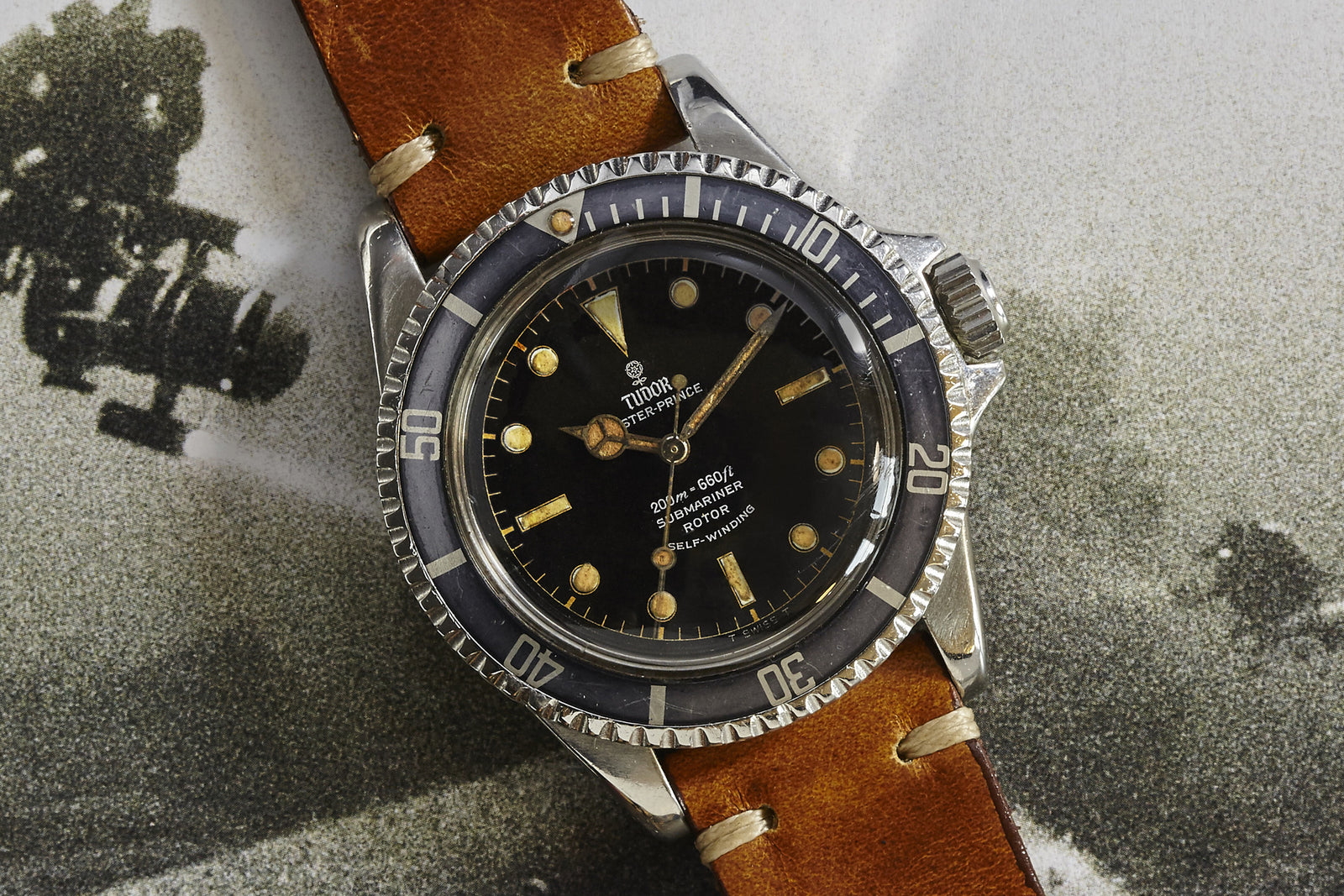 The Tudor Submariner Gilt Dial wristwatch by Tudor, featuring a black dial with gold hands, luminescent markers, a rotating bezel, and a brown leather strap, is displayed on a textured black-and-white background.