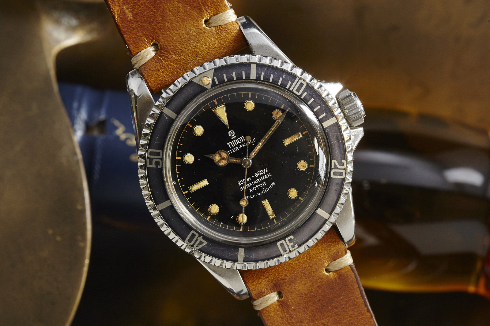 A Tudor Submariner Gilt Dial wristwatch by Tudor features a black dial with round markers, rotating bezel, and brown leather strap, shown in close-up against a blurred background.
