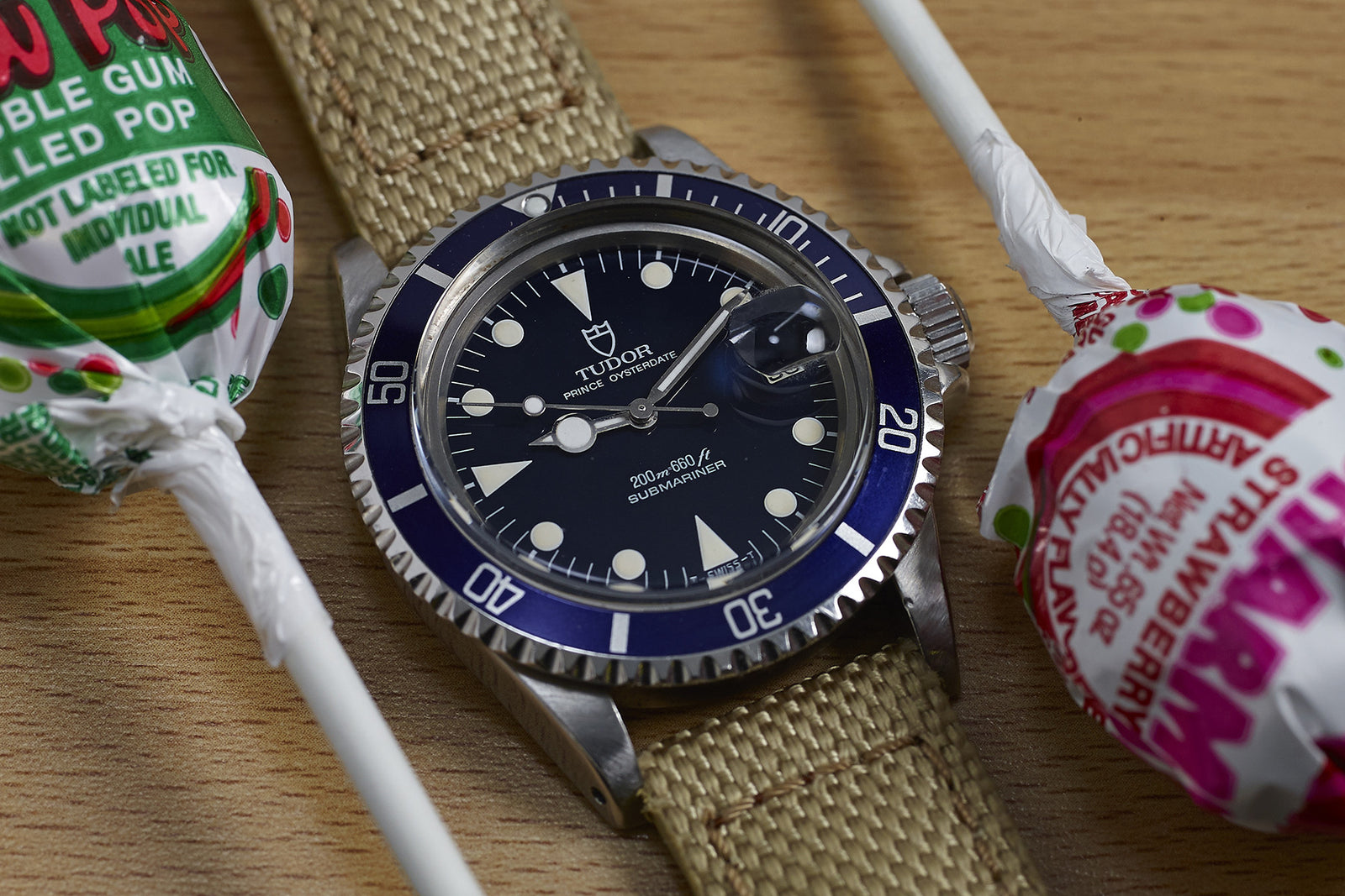 A Tudor Submariner "Lollipop" Blue Dial Ref. 76100 wristwatch with a beige strap sits on wood between two wrapped, colorful Tootsie Pop lollipops.