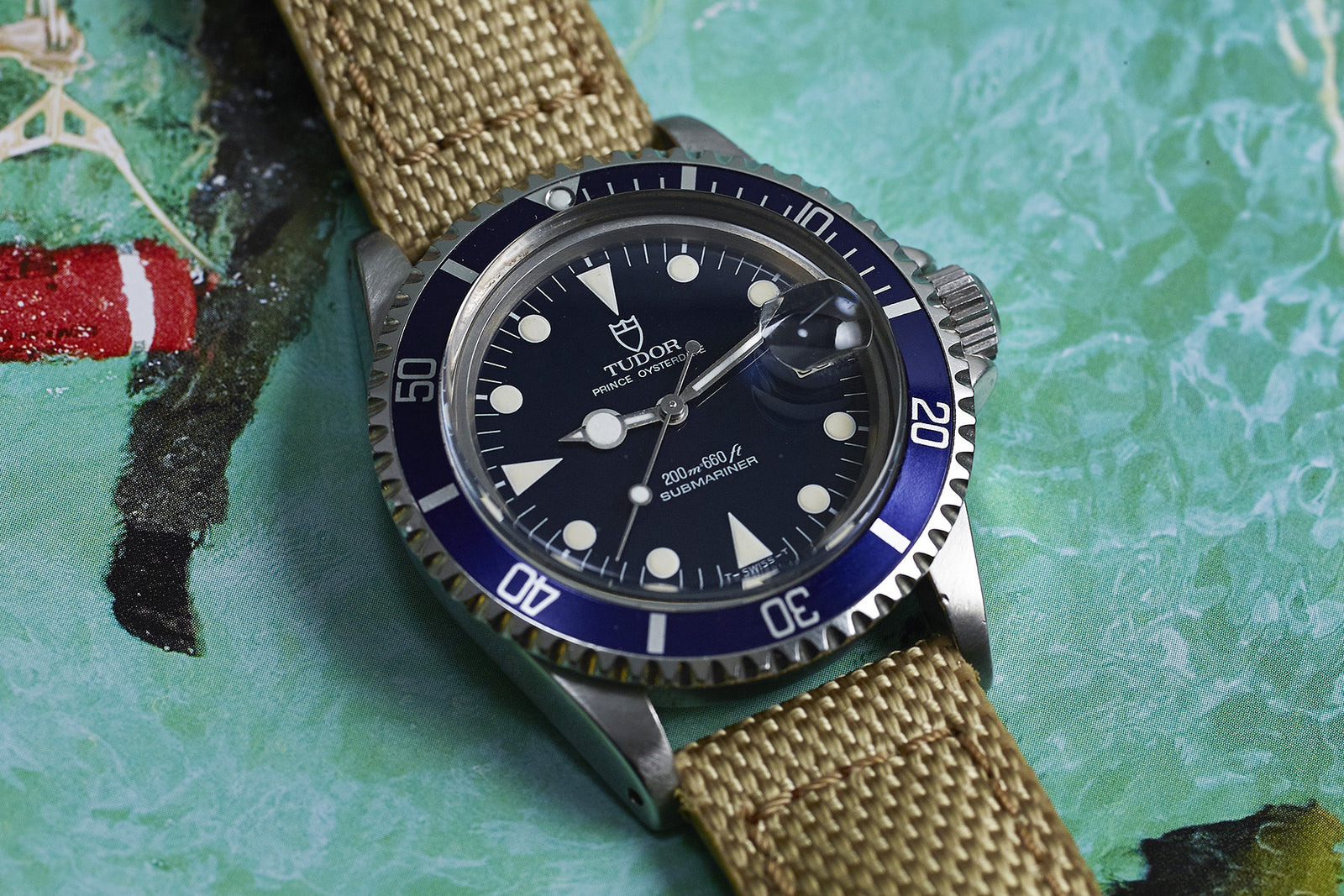 The Tudor Submariner "Lollipop" Blue Dial Ref. 76100, featuring a blue bezel, white markers, and a tan fabric strap, is displayed on a vibrant water-themed background.