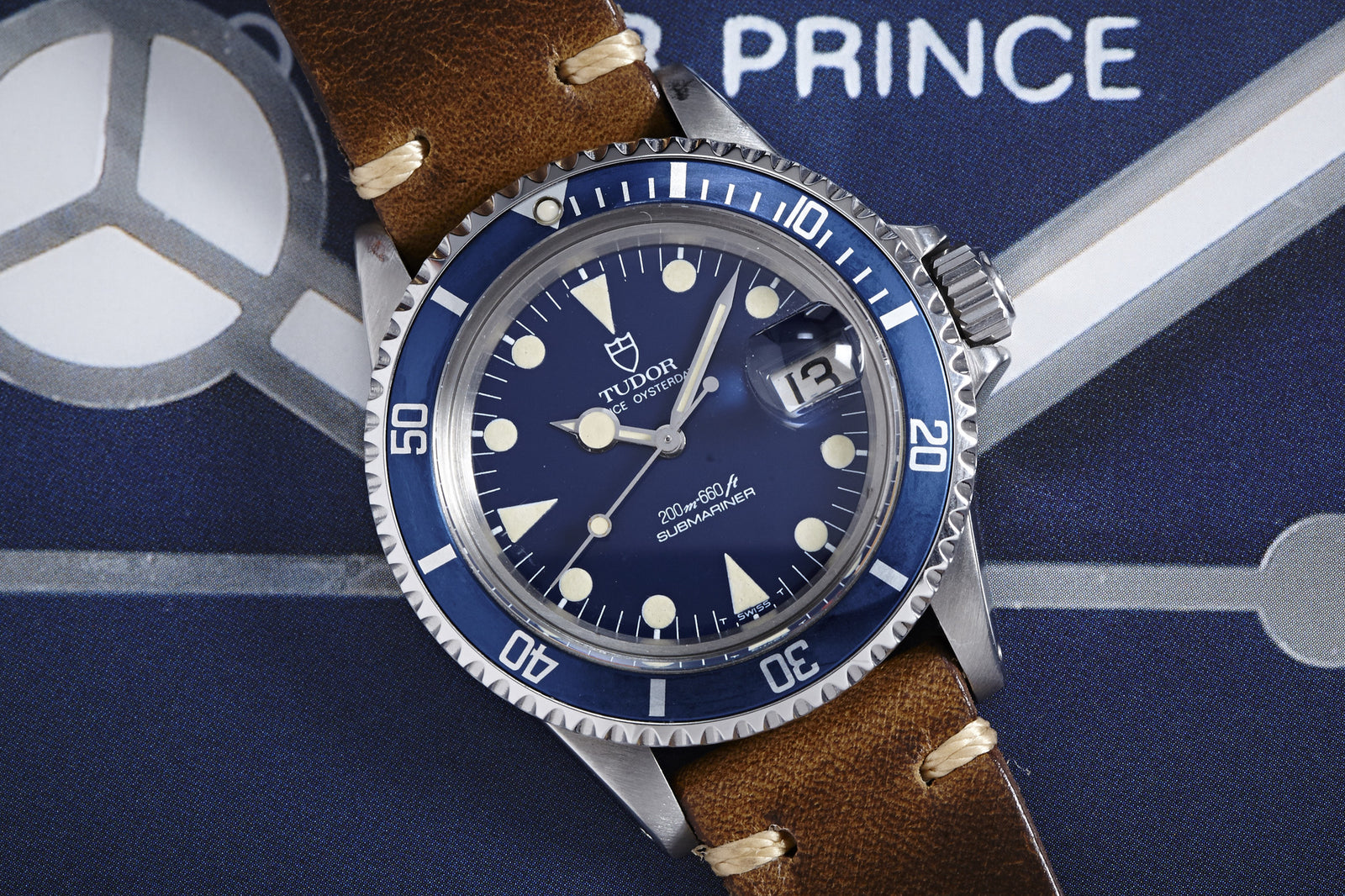 The Tudor Submariner "Lollipop" Ref. 76100 by Tudor features a blue bezel and dial, cream hour markers, and a brown leather strap, shown resting on blue fabric with white text and graphics. Description coming soon.