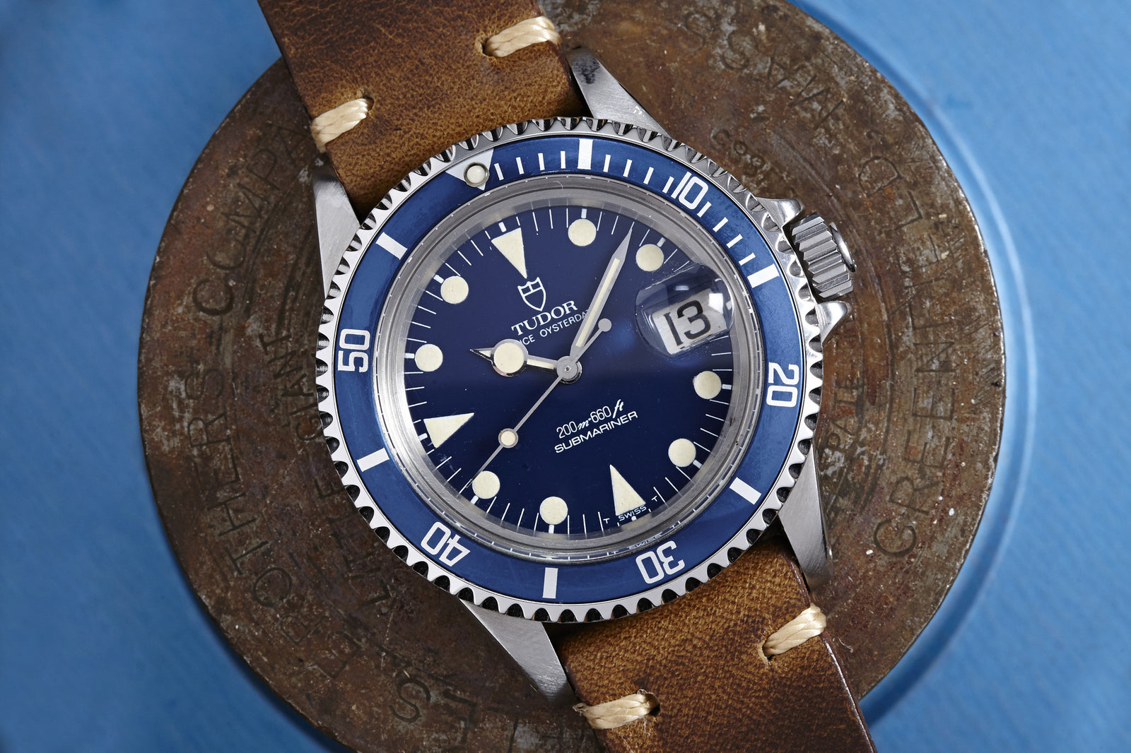 A Tudor Submariner "Lollipop" Ref. 76100 wristwatch by Tudor, featuring a blue dial and bezel, large white hour markers, and date display at 3 o’clock on a brown leather strap, is displayed on a round metal object against a blue background.