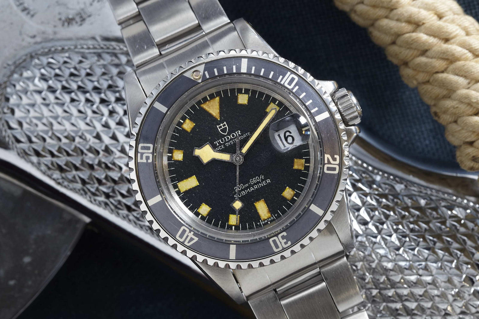 A close-up of a Tudor Submariner by Tudor with a stainless steel band, black bezel, and aged hour markers, shown on a textured metal surface beside a coiled rope.