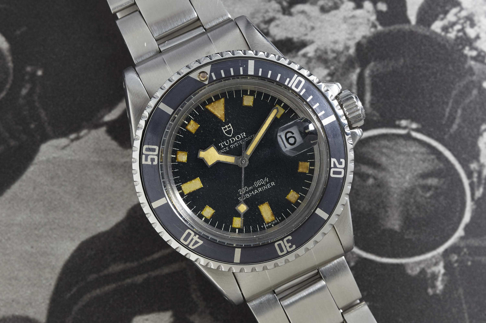 A close-up of a Tudor Submariner by Tudor shows its black bezel, black dial with date window, and stainless steel bracelet, set against a black-and-white background.