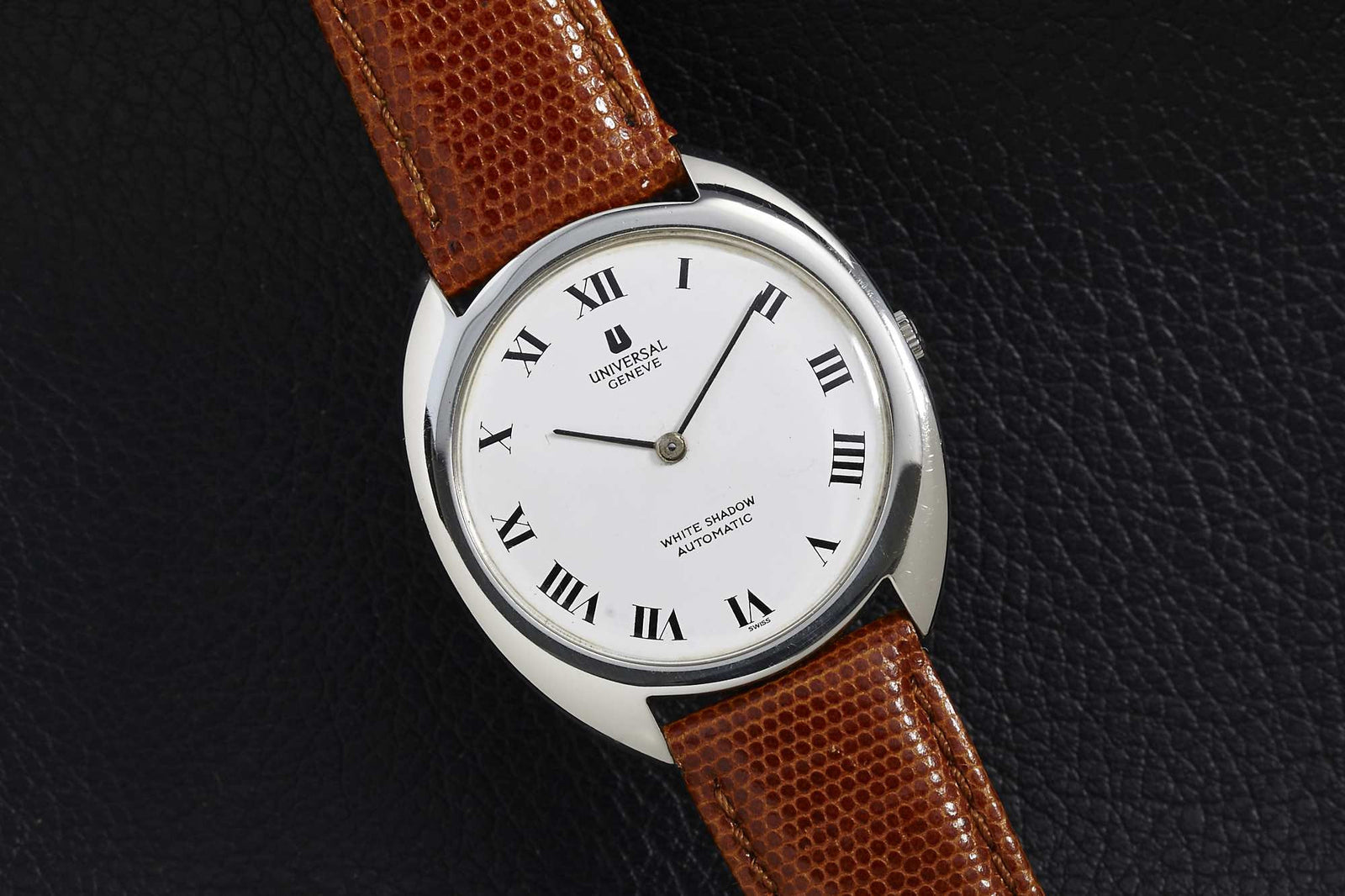 The Universal Geneve White Shadow wristwatch features a white dial with black Roman numerals, a brown textured leather strap, and a polished silver steel case—showcasing vintage elegance on a black surface.