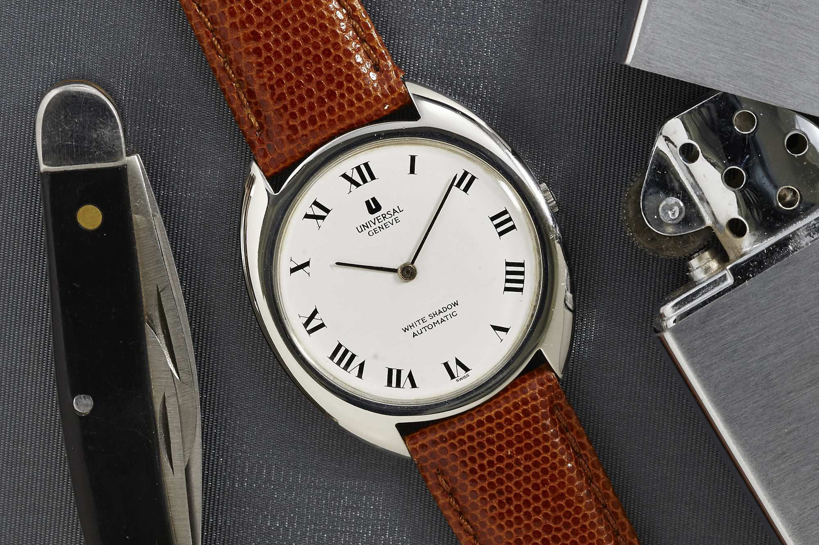 The Universal Geneve White Shadow, a slim automatic dress watch with brown leather strap and white Roman numeral dial, rests between a folding pocket knife and silver lighter on a gray textured surface.