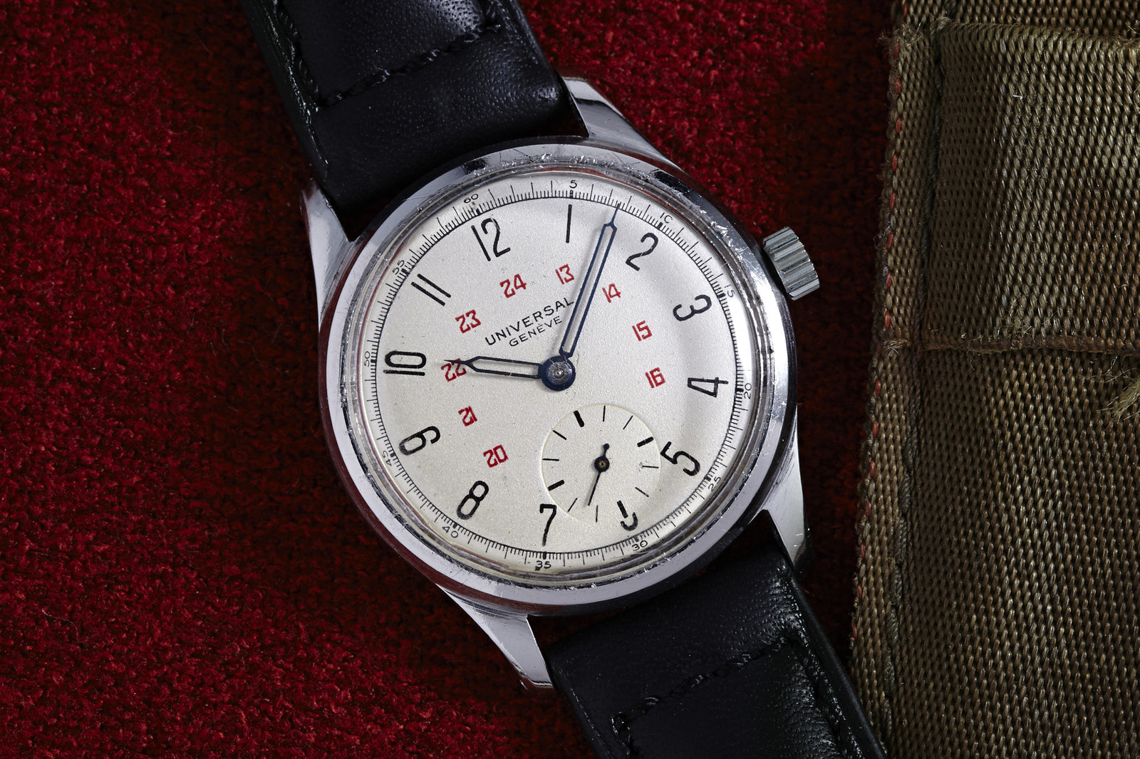 A Universal Geneve Manual Wind Sub Seconds Arabic Dial wristwatch with a silver dial, black numerals, red 24-hour markers, and a black leather strap rests on a red textured surface next to a khaki fabric item. Detailed description coming soon.