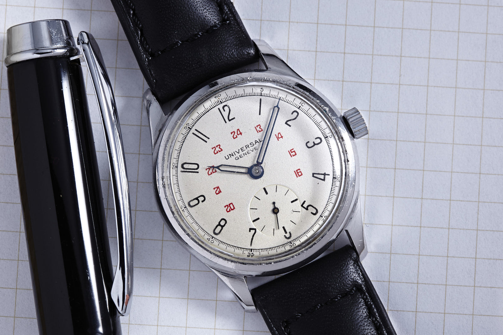 The Universal Geneve Manual Wind Sub Seconds Arabic Dial, featuring a black leather strap and silver case with 12-hour and 24-hour markings, rests beside a black pen on graph paper. More details coming soon.