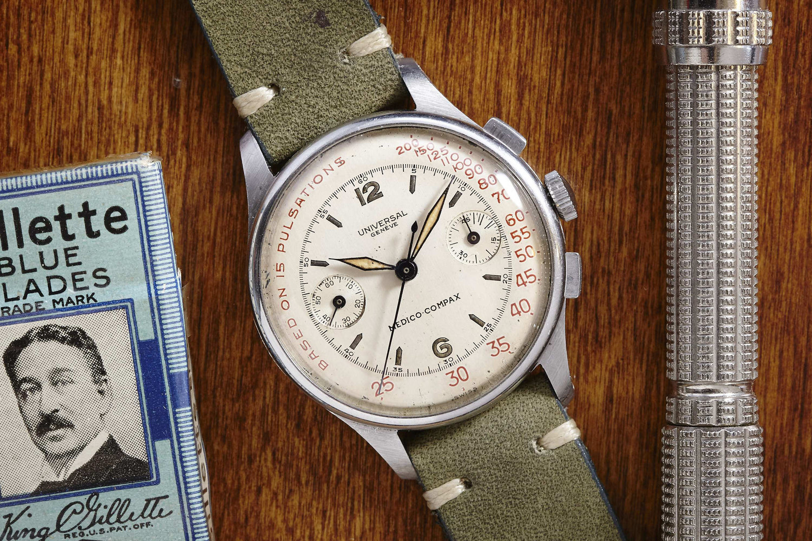 A vintage Universal Geneve Medico-Compax chronograph with a cream dial and green strap sits on wood next to a safety razor and a Gillette Blue Blades box with a man's portrait.