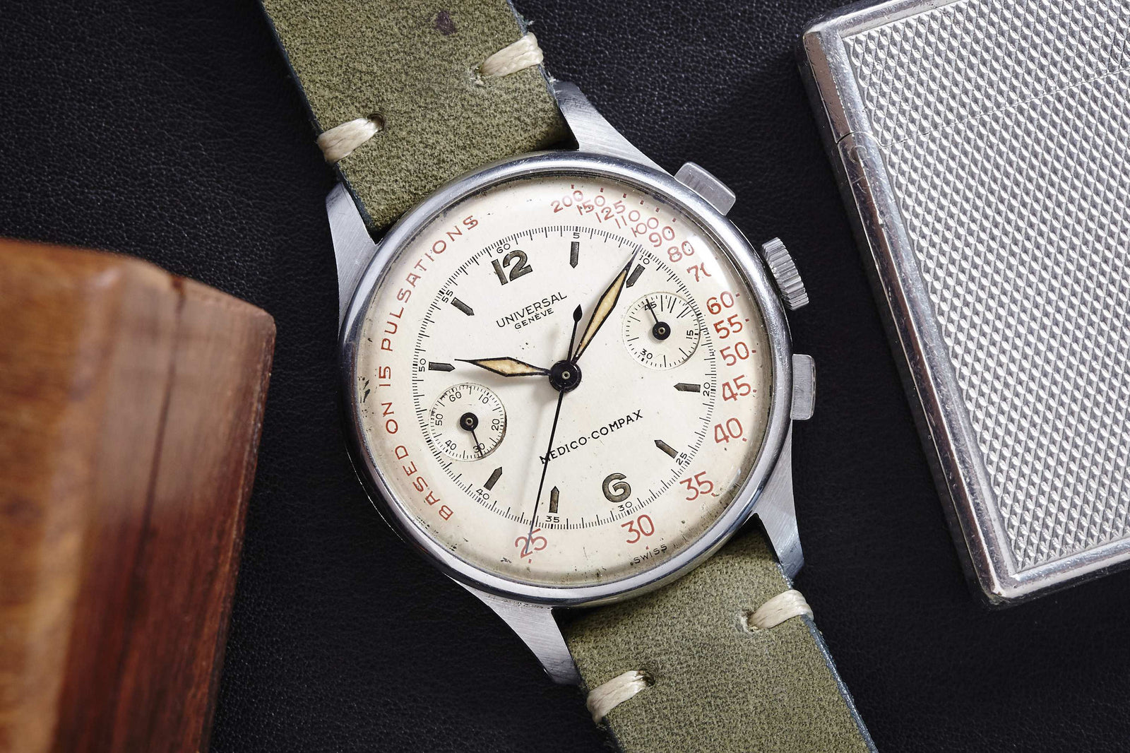 A Universal Genève Medico-Compax chronograph wristwatch with a white dial, two subdials, red pulsations scale, and olive green leather strap rests on a dark surface beside a wooden object and metal case.
