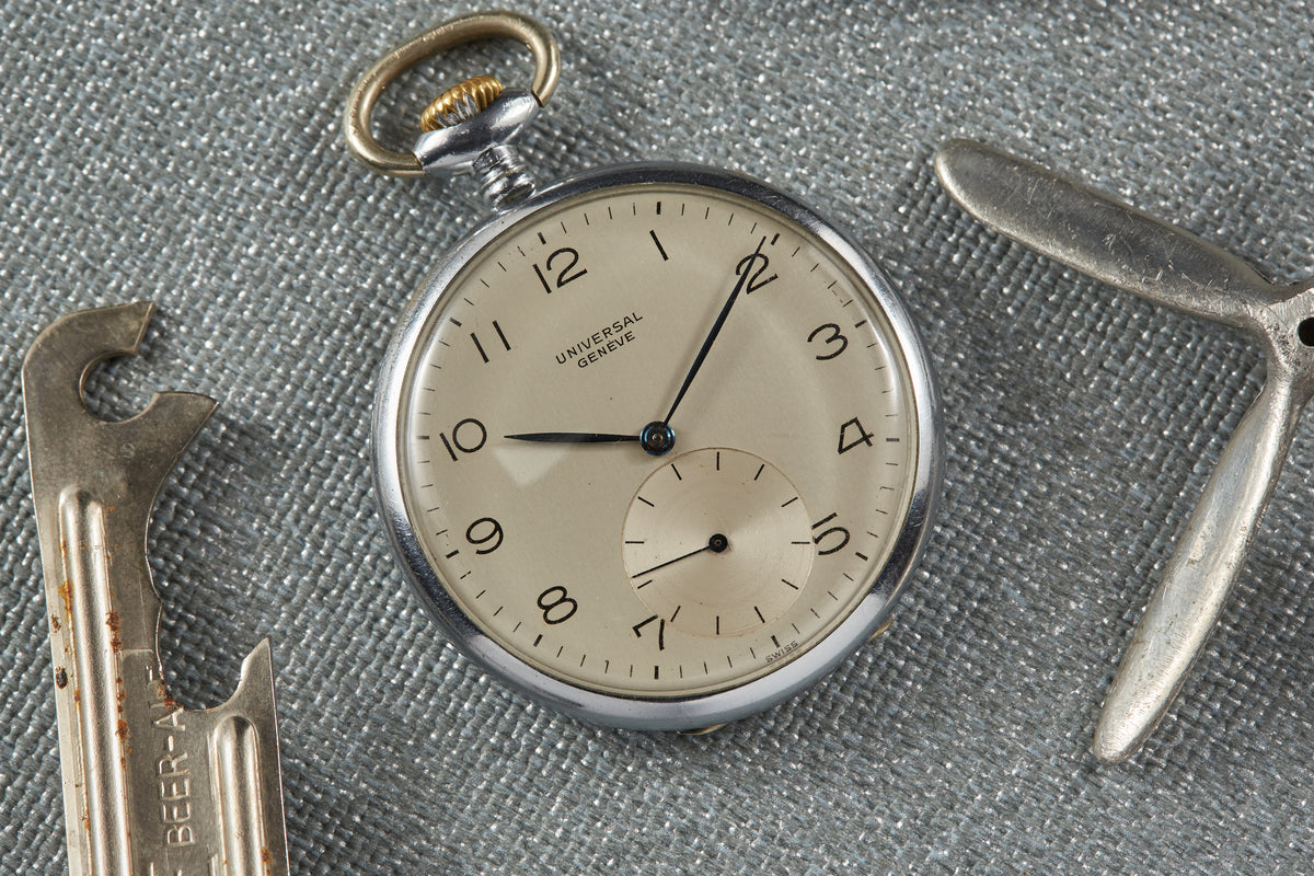 Universal Genève pocket watch with cream dial, Arabic numerals, and subdial, displayed with vintage watch tools on gray fabri