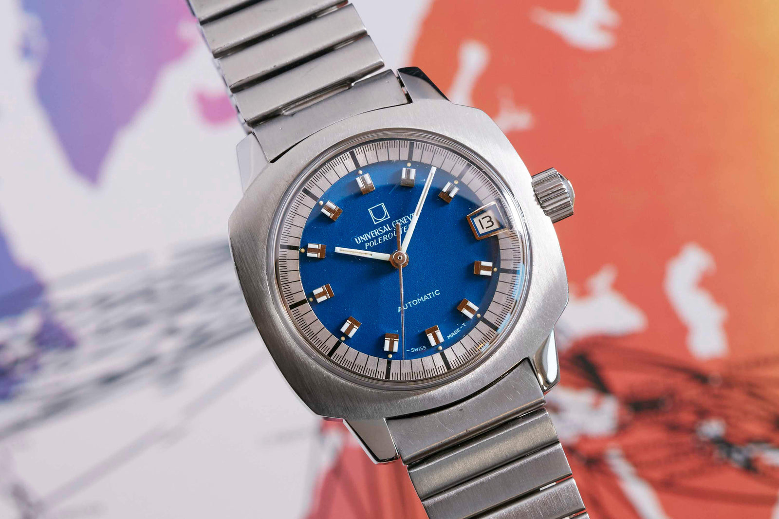 The Universal Genève Polerouter Blue - Cushion Case by Universal Geneve features a stainless steel design, blue dial, silver hour markers, and a date window at 3 o'clock, shown on a metal bracelet against a vibrant blurred background.