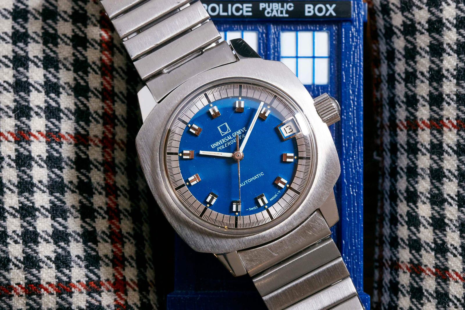 A Universal Genève Polerouter Blue - Cushion Case wristwatch by Universal Geneve with a blue dial and date window sits atop a blue TARDIS model from Doctor Who, set against black, white, and red houndstooth fabric.