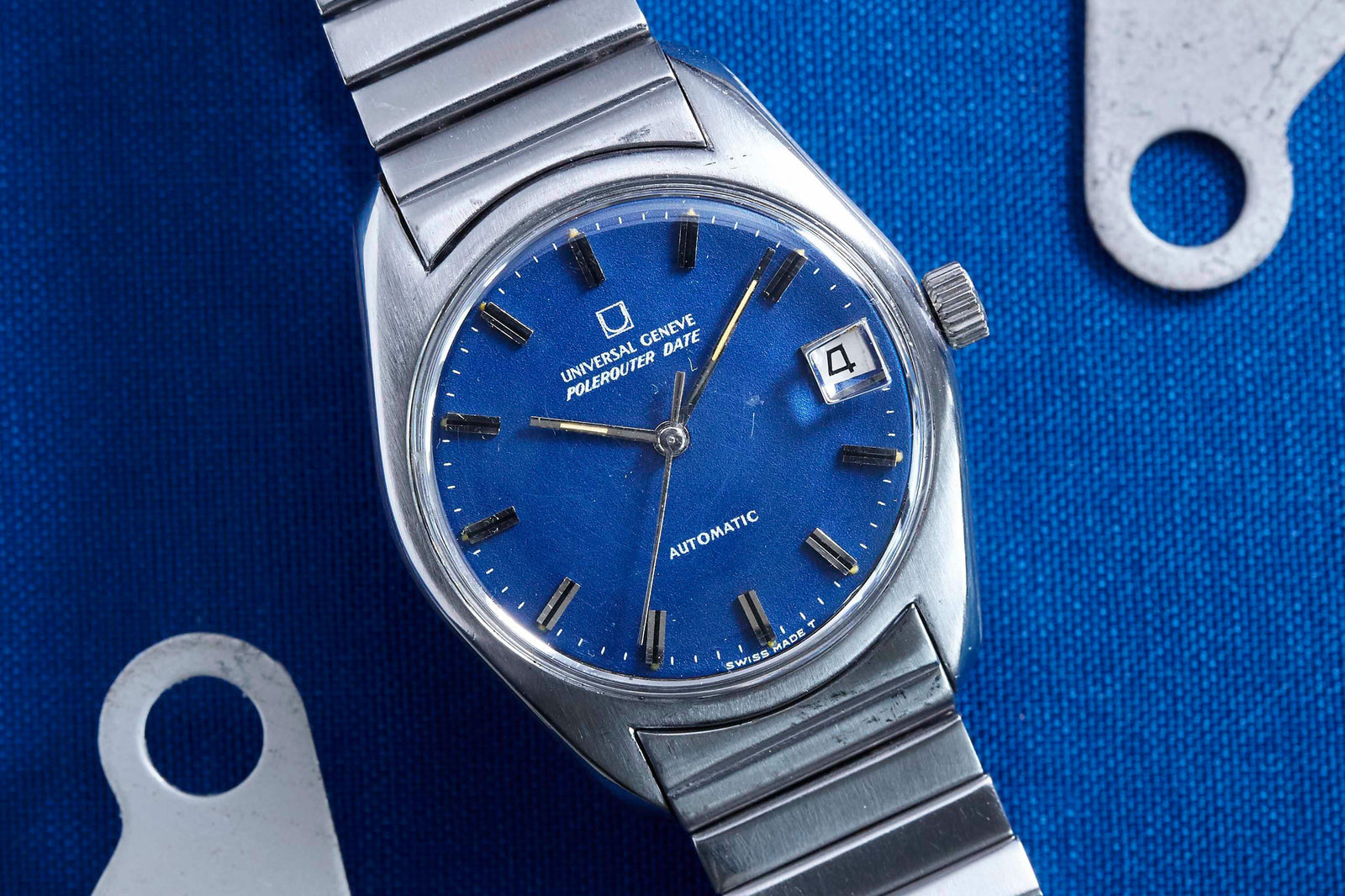 The Universal Genève Polerouter Date by Universal Geneve, designed by Gerald Genta, features a blue dial with silver hour markers and a date window at 3 o’clock. Displayed on blue fabric with metallic parts, it's an icon among luxury stainless steel sports watches.