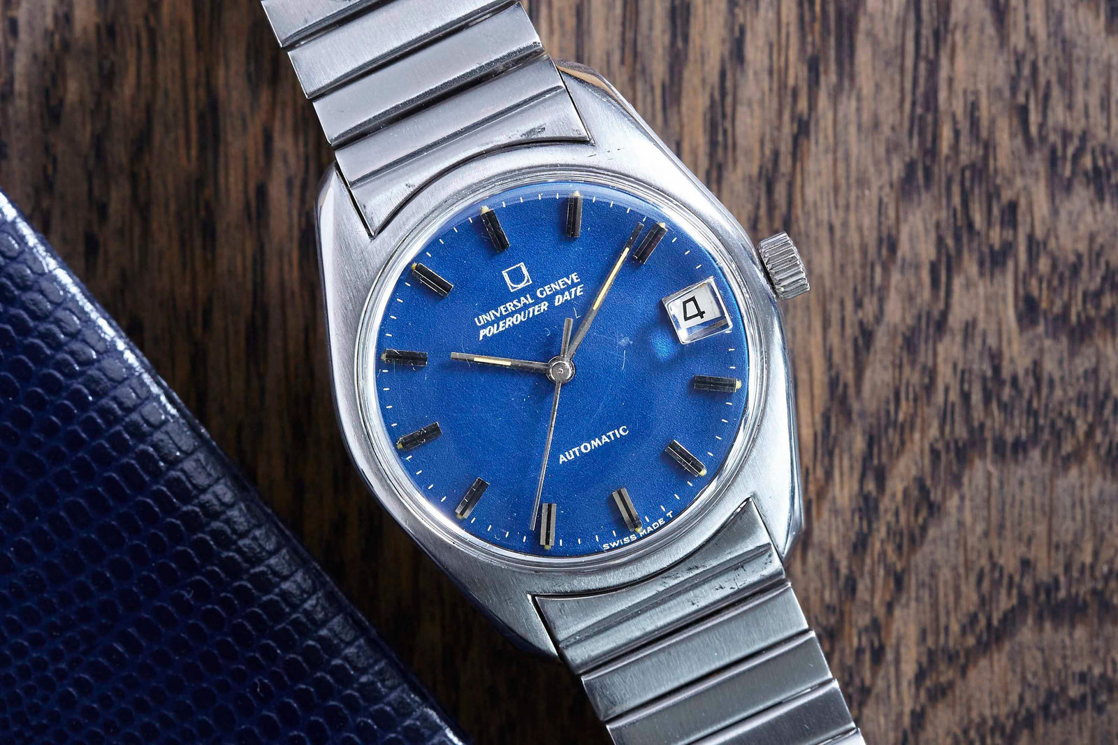The Universal Geneve Polerouter Date wristwatch features a blue dial, silver hour markers, a date window at 3 o’clock, and a silver bracelet, embodying the timeless appeal of classic luxury stainless steel sports watches.