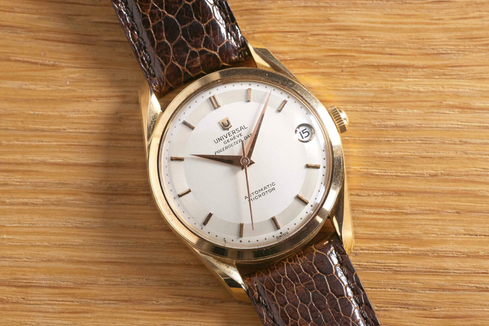 The Universal Genève Polerouter Date by Universal Geneve features a gold case, white dial, brown leather strap, and date window at 3 o’clock—a classic vintage dress watch shown elegantly on a wooden surface.