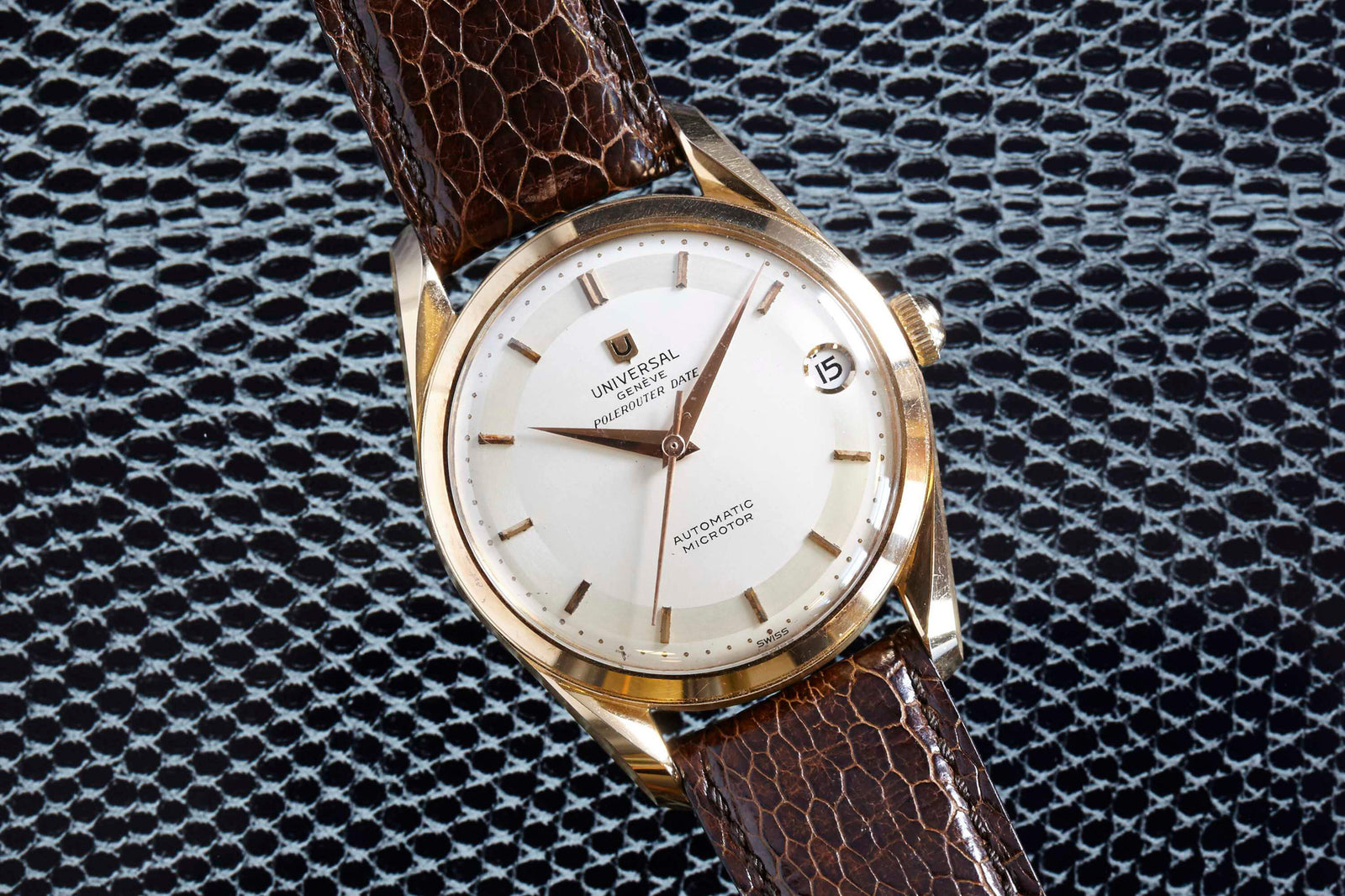The Universal Genève Polerouter Date, a vintage dress watch by Universal Geneve, features a gold case, white dial with gold hour markers, date window at 3 o’clock, and brown leather strap, shown on a textured dark surface.